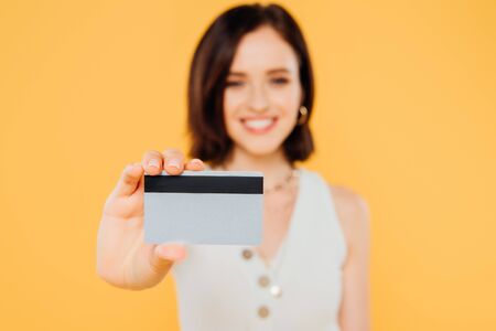 selective focus of smiling elegant girl holding credit card isolated on yellowの写真素材