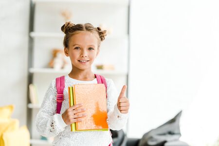 happy schoolgirl holding books and showing thumb up at homeの写真素材