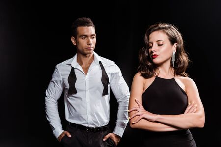 handsome mixed race man standing with hands in pockets near woman with crossed arms isolated on blackの写真素材