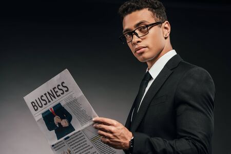 side view of african american businessman in glasses and suit reading business newspaper on dark...