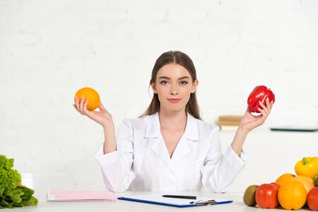 Photo pour front view of dietitian in white coat holding orange and bell pepper at workplace - image libre de droit