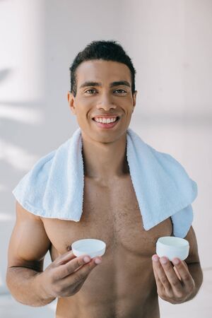 mixed race young man with blue towel on shoulders holding opener container with cosmetic cream while looking at cameraの写真素材