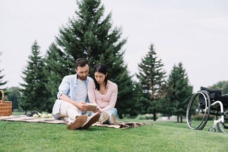 handsome man with disabled girlfriend reading book while sitting on blanket in park