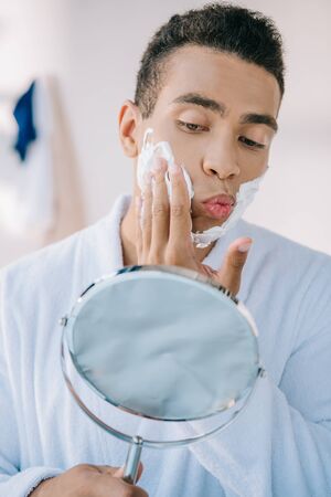 handsome young man in bathrobe applying shaving cream on face while holding mirrorの写真素材