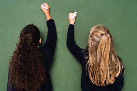 back view of two multicultural schoolgirls writing on chalkboard with chalks