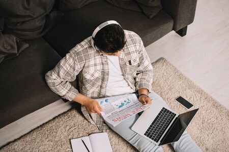 overhead view of young man reading fake news newspaper while sitting on floor with laptop