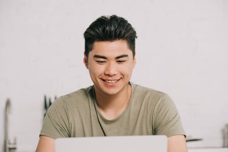 cheerful asian man using laptop while sitting at kitchen tableの写真素材