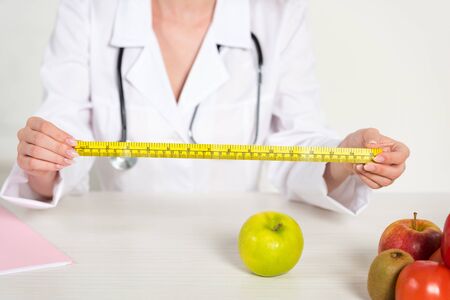 cropped view of dietitian in white coat holding measure tape and fresh food on tableの写真素材