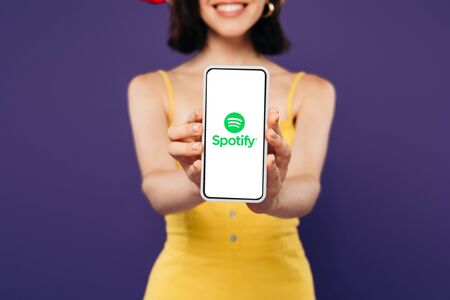 KYIV, UKRAINE - JULY 3, 2019: selective focus of smiling girl presenting smartphone with spotify app isolated on purpleのeditorial素材