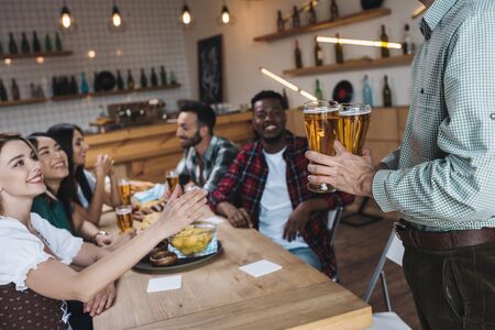 cropped view of man holding glasses of light beer near multicultural friends in pub