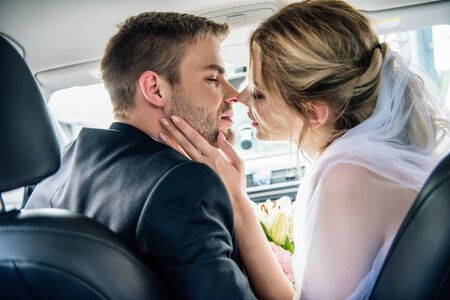 back view of attractive bride in bridal veil and bridegroom kissing in carの写真素材