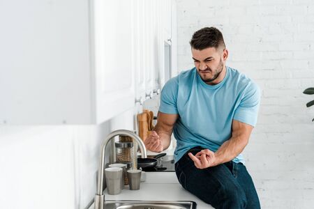 selective focus of emotional man showing middle fingers and looking at faucet in kitchenの写真素材