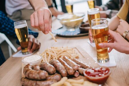 cropped view of man taking french fries and fried sausages on wooden tray in pubの写真素材