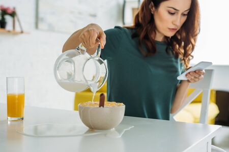 woman overfilling bowl of flakes with water while sitting at kitchen table and using smartphone