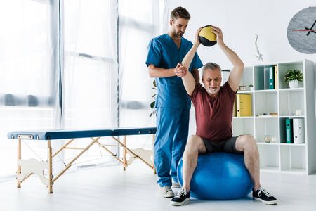 handsome doctor standing near mature man exercising on fitness ballの写真素材