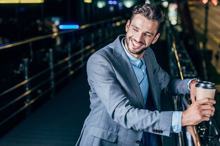 handsome businessman in formal wear smiling and holding paper cup in night city