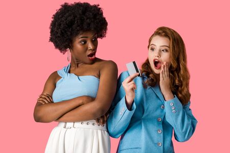 surprised redhead girl holding credit card near shocked african american woman standing with crossed arms isolated on pinkの写真素材