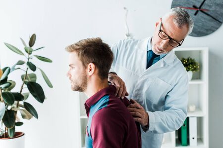 bearded chiropractor in white coat and glasses touching back of handsome patient in clinicの写真素材
