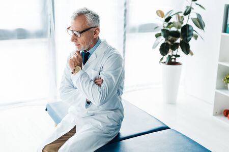pensive bearded doctor in glasses and white coat sitting on massage table and touching faceの写真素材