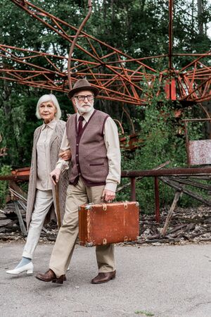 senior man in glasses holding suitcase while walking with wife near abandoned carouselの写真素材