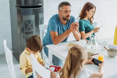 parents with daughter and som using smartphones while sitting in kitchen at table with served...