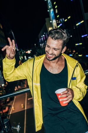handsome man in yellow jacket showing peace sign and holding plastic cup in night city