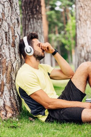 sportsman in headphones covering face while holding sport bottle near tree trunkの写真素材