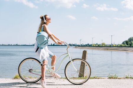 side view of girl with backpack biking and listening music in headphones near river in summerの写真素材