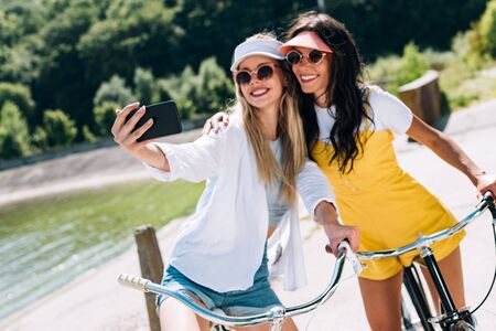selective focus of blonde and brunette girls riding bikes and taking selfie in summerの写真素材