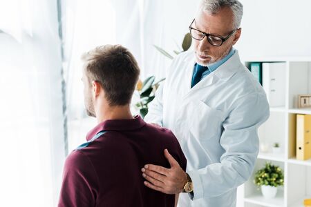 doctor in glasses and white coat touching back of man in clinicの写真素材
