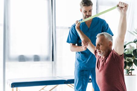 doctor standing near middle aged man exercising with resistance band