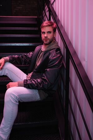 pensive bearded young man in jacket sitting on stairs