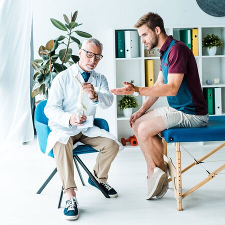 handsome bearded man gesturing while looking at spine model near doctor in glassesの写真素材