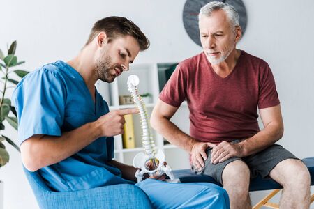 handsome doctor pointing with finger at spine model near patient in clinic