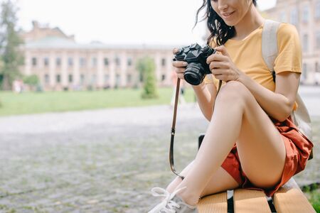 cropped view of young woman holding digital camera while sitting on bench near universityの写真素材