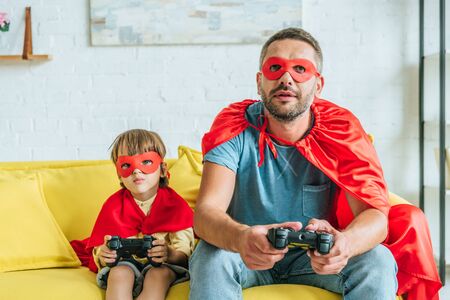KYIV, UKRAINE - JULY 5, 2019: Father and son in costumes of superheroes playing video game while sitting on sofa at homeの写真素材