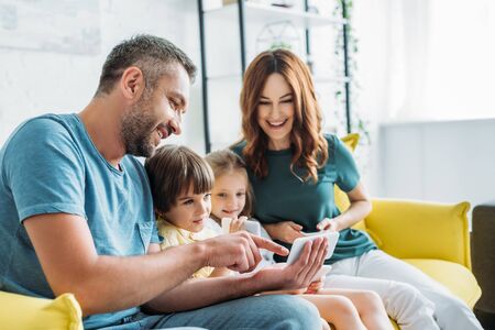 happy father pointing with finger while showing smartphone to children near smiling wifeの写真素材