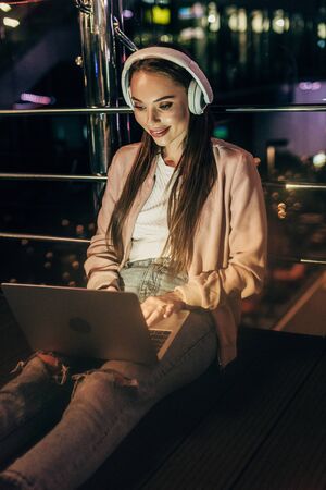 smiling woman in pink jacket with headphones using laptop in night cityの写真素材