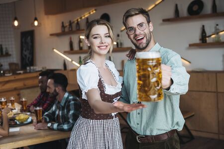 selective focus of handsome young man holding mug of light beer while standing near beautiful waitress in traditional german costumeの写真素材