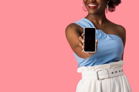 cropped view of cheerful african american girl holding smartphone with blank screen isolated on pinkの写真素材