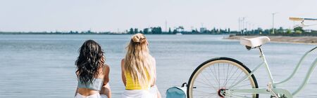back view of blonde and brunette friends sitting near bike and river in summer, panoramic shot