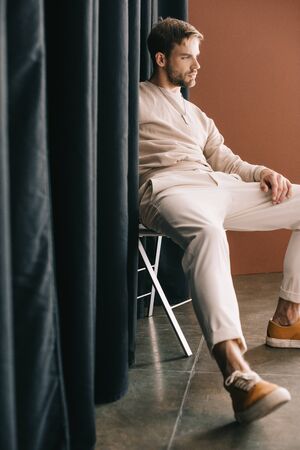 pensive bearded man in casual outfit sitting on chair near curtainの写真素材