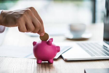 cropped view of businessman putting coin into pink piggy bank near laptopの写真素材