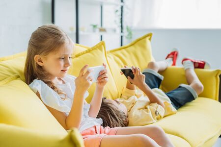 adorable child sitting with smartphone near brother lying on sofa and using smartphone at homeの写真素材
