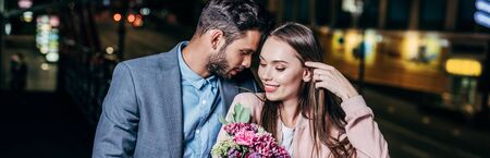 panoramic shot of handsome businessman giving bouquet and kissing attractive woman in night cityの写真素材