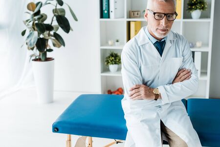 happy bearded doctor in glasses and white coat sitting on massage table with crossed armsの写真素材