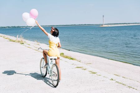 back view of brunette girl riding bike with balloons near riverの写真素材