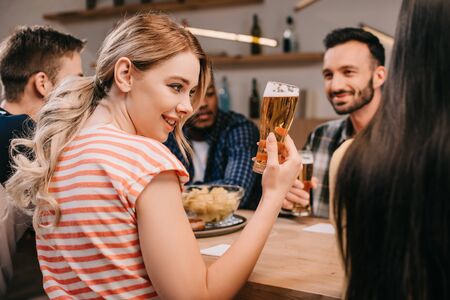 selective focus of smiling young woman holding glass of beer while sitting with multicultural friends in pubの写真素材