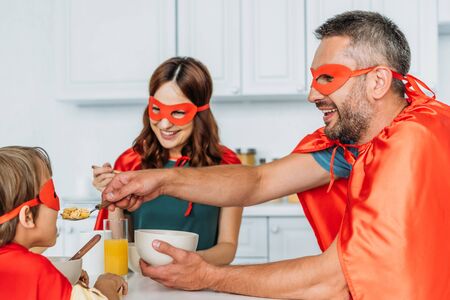 family in superheroes costumes having breakfast while father feeding son with flakesの写真素材