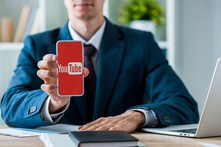 KYIV, UKRAINE - JULY 8, 2019: selective focus of man holding smartphone with youtube app on screen near laptop in officeのeditorial素材